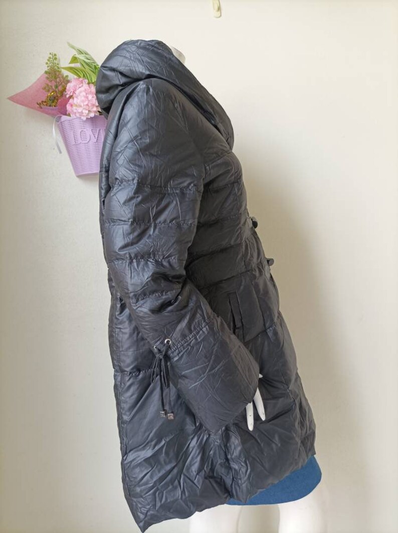 May include: A dark grey puffer coat with a hood, displayed on a mannequin. The coat features a quilted design, a cinched waist, and drawstring details on the sleeves. The coat is long, extending to the mid-thigh.