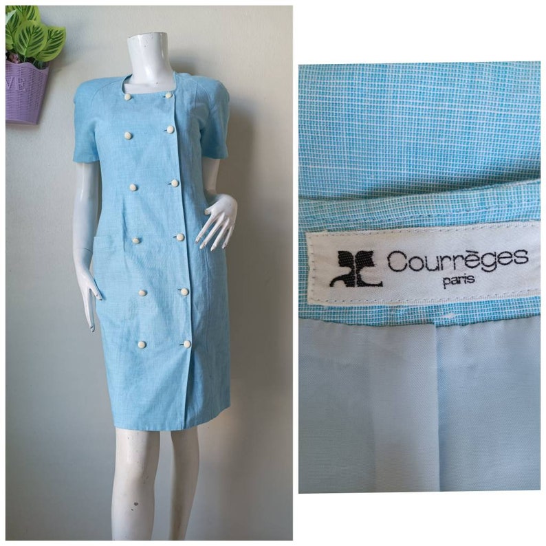 May include: A light blue dress with a double breasted button closure and short sleeves. The dress has a white label with the text "Courr&egrave;ges Paris" on the bottom.