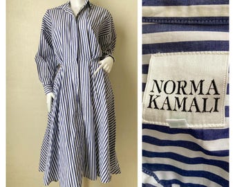 May include: A midi-length shirt dress featuring a vertical blue and white striped pattern, a collared neckline, long sleeves, and a cinched waist. A white fabric label with the black text NORMA KAMALI is stitched onto the striped fabric.