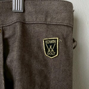 May include: Close-up of brown herringbone golf pants with the "EDWIN GOLF" logo patch. The pants are made of a textured fabric and have a metal clasp at the top. The pants are hanging from a metal hanger.