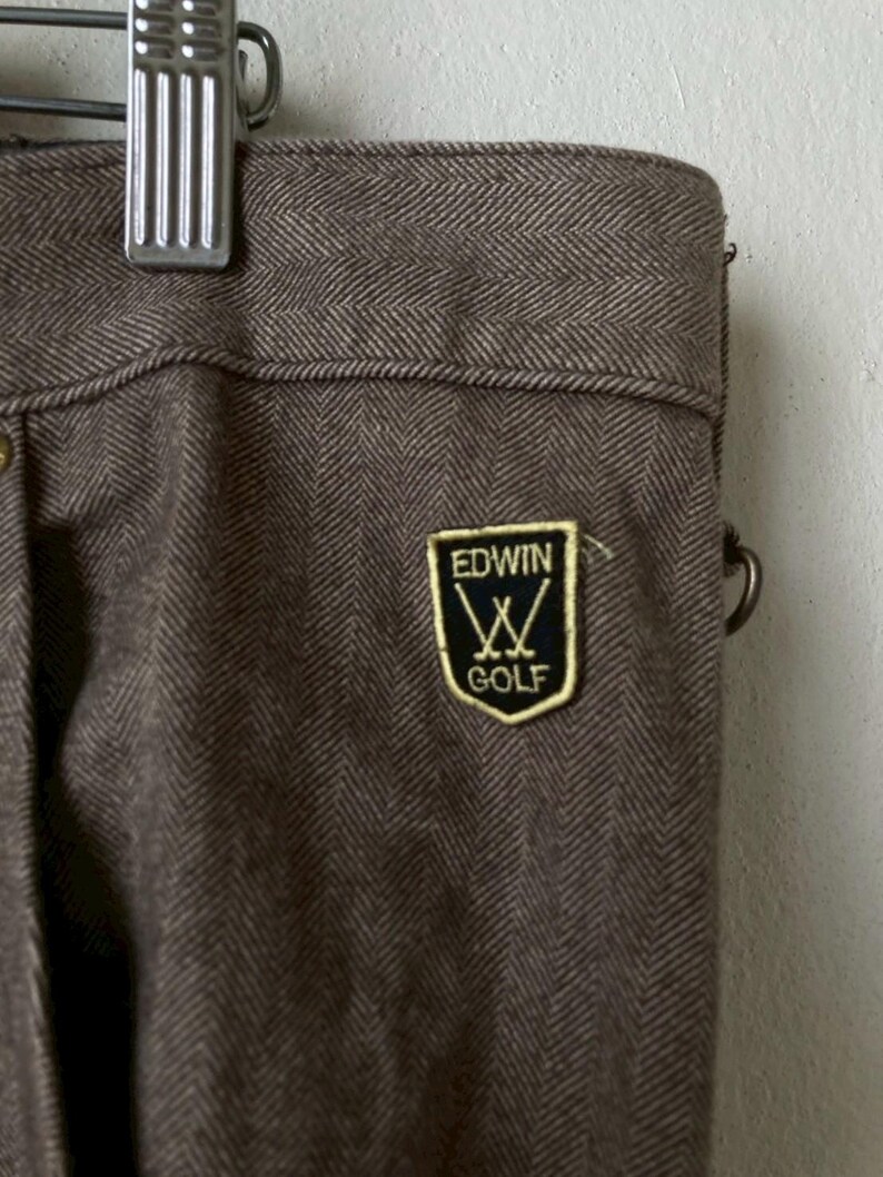 May include: Close-up of brown herringbone golf pants with the "EDWIN GOLF" logo patch. The pants are made of a textured fabric and have a metal clasp at the top. The pants are hanging from a metal hanger.