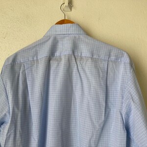 May include: A light blue button-down shirt with a fine white grid pattern. The shirt is hanging on a wooden hanger.