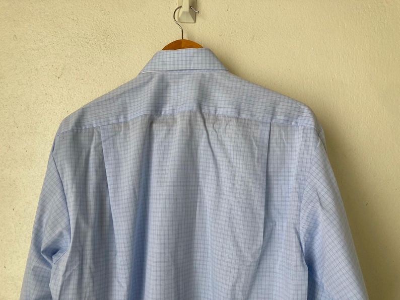 May include: A light blue button-down shirt with a fine white grid pattern. The shirt is hanging on a wooden hanger.