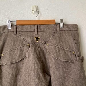 May include: Brown denim pants with unique pocket design and gold-colored rivets. The pants feature a small, triangular logo on the back pocket. The pants are hanging on a wooden hanger.