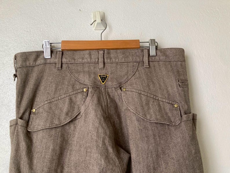 May include: Brown denim pants with unique pocket design and gold-colored rivets. The pants feature a small, triangular logo on the back pocket. The pants are hanging on a wooden hanger.