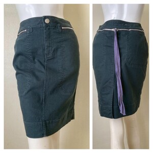 May include: Dark teal pencil skirt with silver zipper details and a button closure. The skirt has a purple tie detail at the back and features pockets. The skirt is displayed on a mannequin.