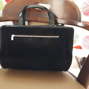K&ouml;nnte beinhalten: Eine schwarze Cord-Tragetasche mit einer Rei&szlig;verschlusstasche und einem silbernen Rei&szlig;verschluss. Die Tasche hat einen braunen Ledergriff.