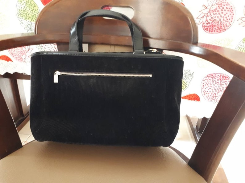 K&ouml;nnte beinhalten: Eine schwarze Cord-Tragetasche mit einer Rei&szlig;verschlusstasche und einem silbernen Rei&szlig;verschluss. Die Tasche hat einen braunen Ledergriff.