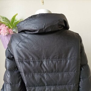 May include: A dark grey puffer jacket with a high collar. The jacket features horizontal stitching and a shiny finish. The back of the jacket is visible, and it appears to be made of a lightweight material. The jacket is displayed on a white mannequin head.