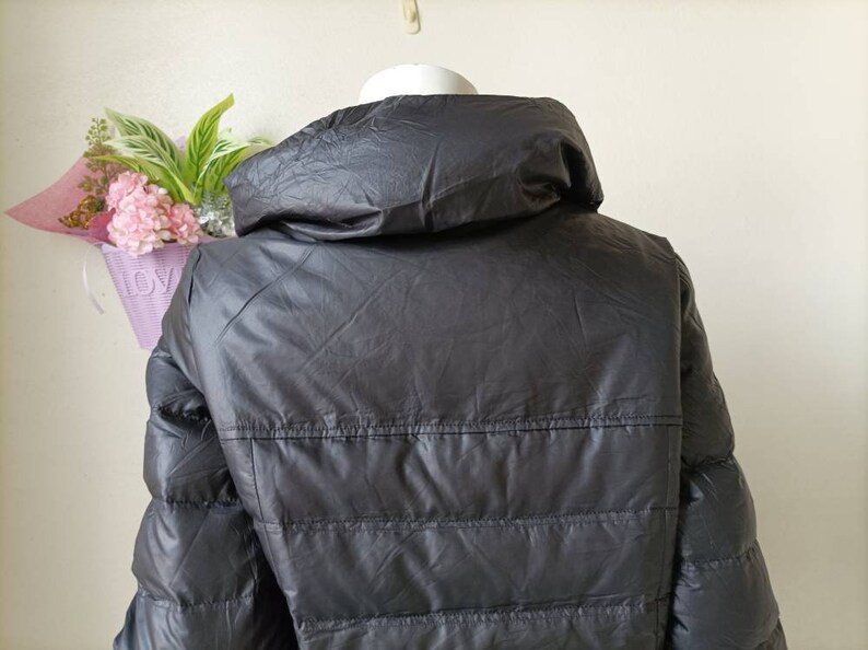 May include: A dark grey puffer jacket with a high collar. The jacket features horizontal stitching and a shiny finish. The back of the jacket is visible, and it appears to be made of a lightweight material. The jacket is displayed on a white mannequin head.