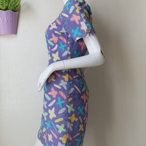 May include: A purple mini dress with a colorful abstract print of lines and crosses. The dress has short sleeves and a scoop neck.