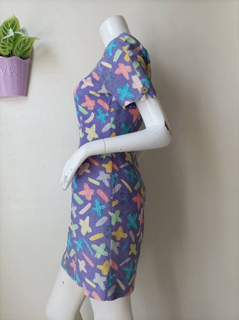 May include: A purple mini dress with a colorful abstract print of lines and crosses. The dress has short sleeves and a scoop neck.