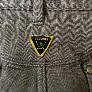 May include: Close-up of gray herringbone-patterned pants. The pants feature a yellow and black Edwin logo patch. The fabric has a textured appearance, and the stitching is visible. The pants have a button closure.