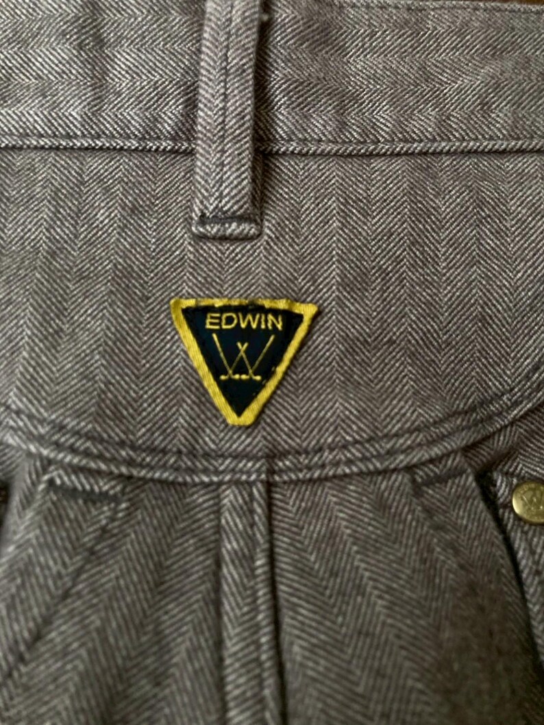 May include: Close-up of gray herringbone-patterned pants. The pants feature a yellow and black Edwin logo patch. The fabric has a textured appearance, and the stitching is visible. The pants have a button closure.