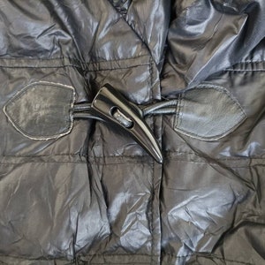 May include: Close-up of a black puffer jacket with a glossy finish. The jacket features a large, black toggle closure and leather-like patches. The stitching is visible, and the fabric has a subtle sheen.