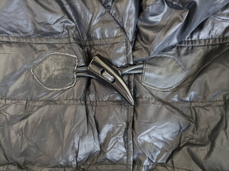May include: Close-up of a black puffer jacket with a glossy finish. The jacket features a large, black toggle closure and leather-like patches. The stitching is visible, and the fabric has a subtle sheen.
