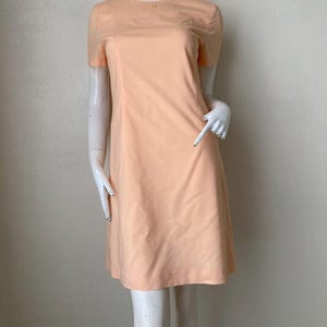 K&ouml;nnte beinhalten: Ein pfirsichfarbenes Kleid mit kurzen &Auml;rmeln. Das Kleid hat ein schlichtes, klassisches Design mit rundem Ausschnitt und einer leicht ausgestellten Silhouette. Das Kleid wird auf einer Schaufensterpuppe pr&auml;sentiert.