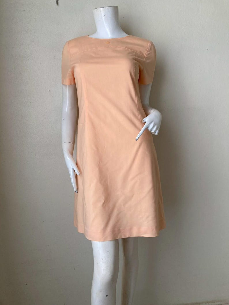 K&ouml;nnte beinhalten: Ein pfirsichfarbenes Kleid mit kurzen &Auml;rmeln. Das Kleid hat ein schlichtes, klassisches Design mit rundem Ausschnitt und einer leicht ausgestellten Silhouette. Das Kleid wird auf einer Schaufensterpuppe pr&auml;sentiert.