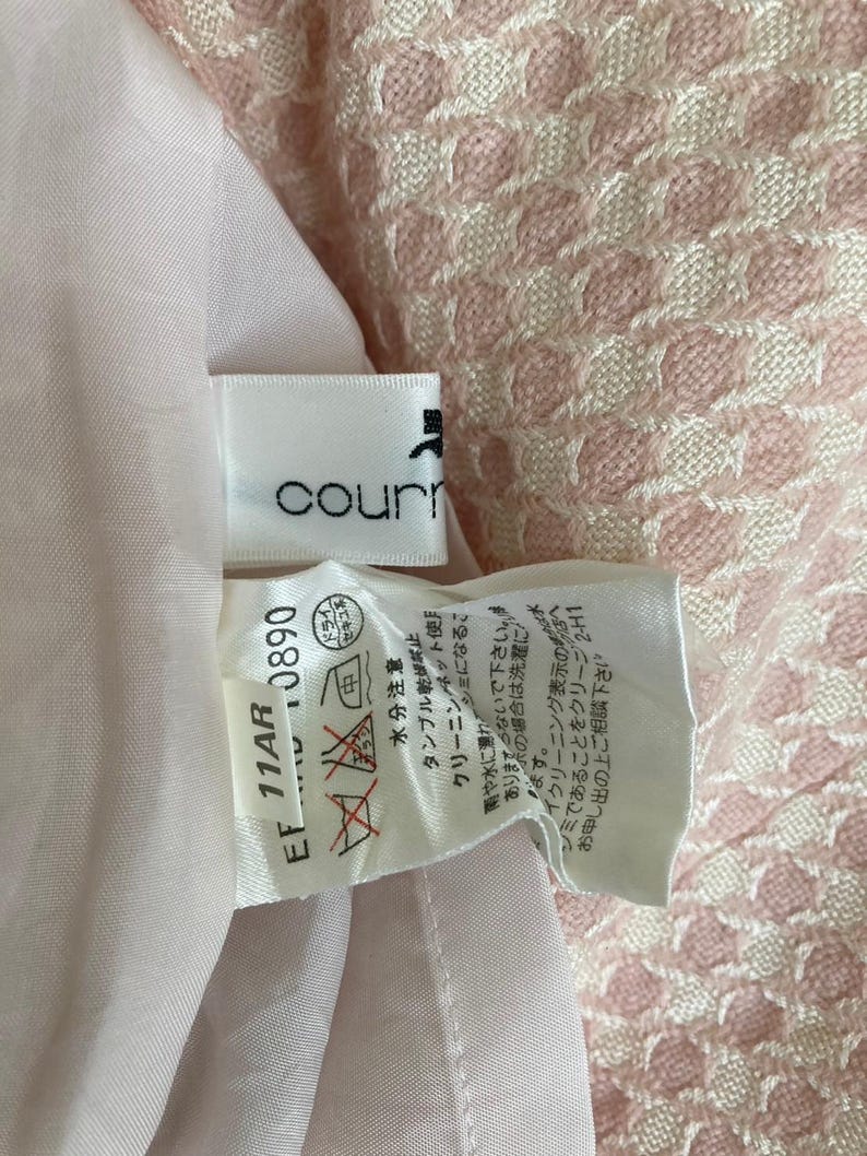 May include: A white tag with black text that reads "courr" and "EF 11AR 0890". The tag has a care label with symbols for washing, drying, and ironing. The tag is attached to a pink and white patterned fabric.