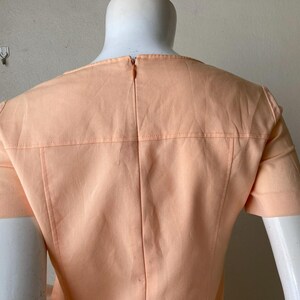 K&ouml;nnte beinhalten: Pfirsichfarbenes Kleid mit kurzen &Auml;rmeln, auf einer Schaufensterpuppe pr&auml;sentiert. Der R&uuml;cken des Kleides hat einen Rei&szlig;verschluss und detaillierte N&auml;hte an Schultern und R&uuml;cken. Das Kleid hat ein schlichtes, elegantes Design.
