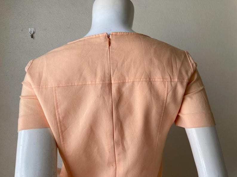K&ouml;nnte beinhalten: Pfirsichfarbenes Kleid mit kurzen &Auml;rmeln, auf einer Schaufensterpuppe pr&auml;sentiert. Der R&uuml;cken des Kleides hat einen Rei&szlig;verschluss und detaillierte N&auml;hte an Schultern und R&uuml;cken. Das Kleid hat ein schlichtes, elegantes Design.