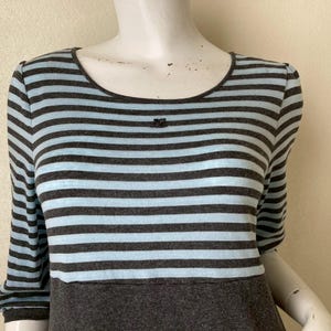 Puede incluir: Vestido a rayas grises y celeste con mangas tres cuartos. El vestido presenta un escote redondo y un corpi&ntilde;o gris oscuro. Un peque&ntilde;o detalle decorativo est&aacute; presente en el escote. El vestido se exhibe en un maniqu&iacute;.