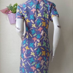 May include: A purple dress with a colorful abstract print. The print features a variety of shapes and colors, including yellow, blue, green, and pink.