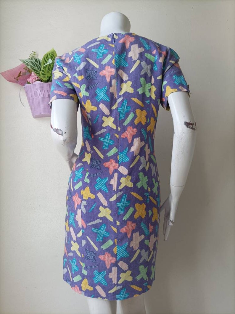 May include: A purple dress with a colorful abstract print. The print features a variety of shapes and colors, including yellow, blue, green, and pink.