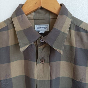 Puede incluir: Una camisa de manga larga a cuadros marrones y verdes con una etiqueta de Burberrys. La camisa tiene un cuello abotonado y un bolsillo en el pecho.