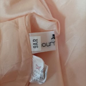 K&ouml;nnte beinhalten: Nahaufnahme des Inneren eines pfirsichfarbenen Kleidungsst&uuml;cks, das ein Etikett mit der Aufschrift "9AR" und einem Markennamen zeigt. Der Stoff ist weich und leicht zerknittert. Ein kleineres Etikett mit japanischem Text ist ebenfalls sichtbar.