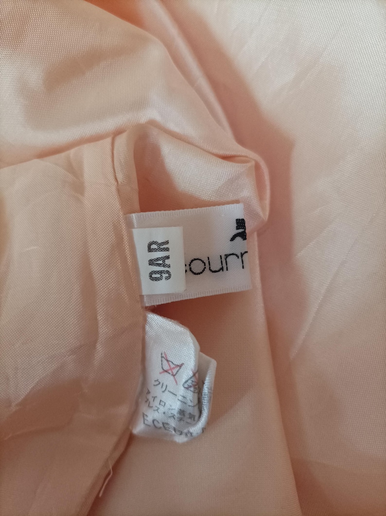 K&ouml;nnte beinhalten: Nahaufnahme des Inneren eines pfirsichfarbenen Kleidungsst&uuml;cks, das ein Etikett mit der Aufschrift "9AR" und einem Markennamen zeigt. Der Stoff ist weich und leicht zerknittert. Ein kleineres Etikett mit japanischem Text ist ebenfalls sichtbar.