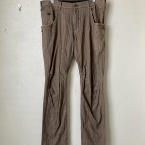 May include: Brown pants with multiple pockets and zippers, hanging on a wooden hanger. The pants have a classic design with a button and zipper closure. The fabric appears to be a durable, woven material. The pants are displayed against a plain white wall.