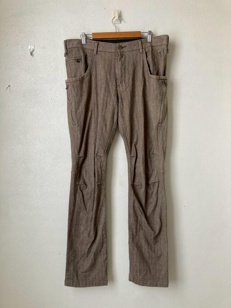 May include: Brown pants with multiple pockets and zippers, hanging on a wooden hanger. The pants have a classic design with a button and zipper closure. The fabric appears to be a durable, woven material. The pants are displayed against a plain white wall.