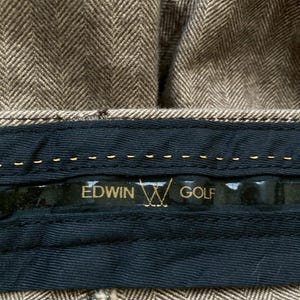 May include: Close-up of brown herringbone fabric with a black label. The label reads "EDWIN GOLF" in gold lettering, with a golf club logo. The fabric appears to be part of a garment, possibly trousers.