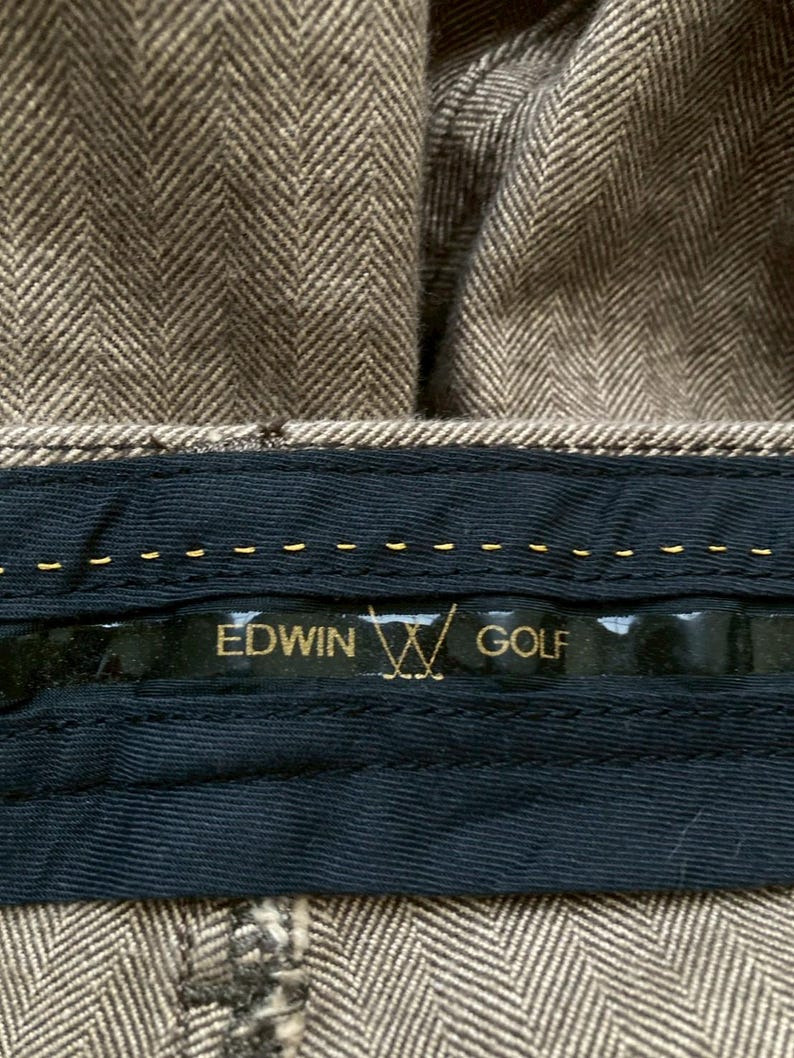 May include: Close-up of brown herringbone fabric with a black label. The label reads "EDWIN GOLF" in gold lettering, with a golf club logo. The fabric appears to be part of a garment, possibly trousers.