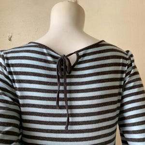 Puede incluir: Una camiseta de manga larga con rayas horizontales en azul claro y gris oscuro. La parte trasera de la camiseta presenta una abertura de ojo de cerradura con un cierre de lazo. Las mangas son largas y la tela parece ser un punto suave.