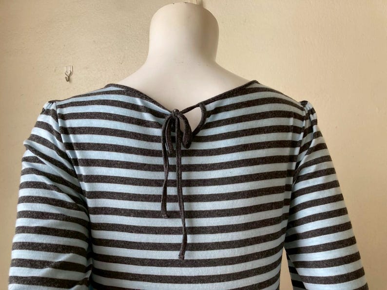 Puede incluir: Una camiseta de manga larga con rayas horizontales en azul claro y gris oscuro. La parte trasera de la camiseta presenta una abertura de ojo de cerradura con un cierre de lazo. Las mangas son largas y la tela parece ser un punto suave.