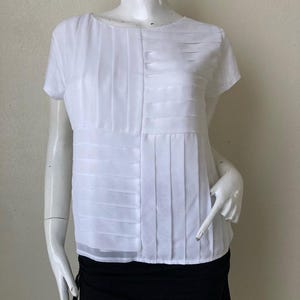 Peut inclure: Blouse blanche &agrave; manches courtes avec d&eacute;tails pliss&eacute;s. Le haut pr&eacute;sente une encolure ronde et un ourlet semi-transparent. La blouse est pr&eacute;sent&eacute;e sur un mannequin, associ&eacute;e &agrave; une jupe noire.