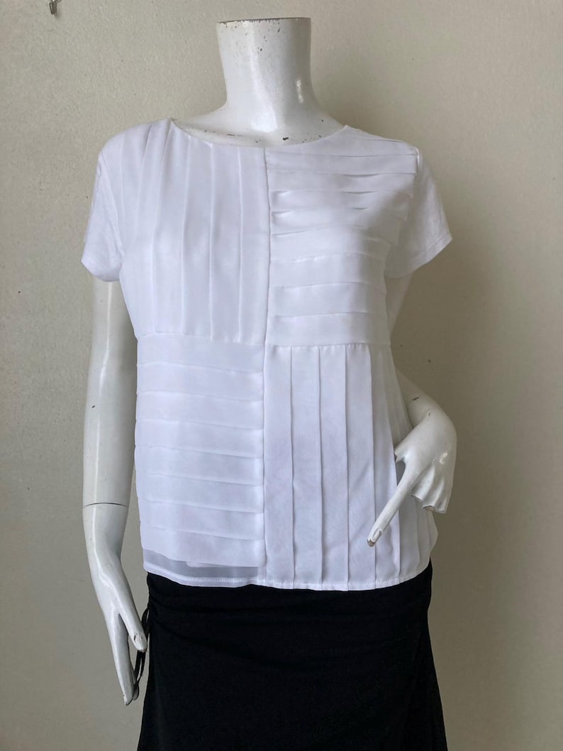 Peut inclure: Blouse blanche &agrave; manches courtes avec d&eacute;tails pliss&eacute;s. Le haut pr&eacute;sente une encolure ronde et un ourlet semi-transparent. La blouse est pr&eacute;sent&eacute;e sur un mannequin, associ&eacute;e &agrave; une jupe noire.