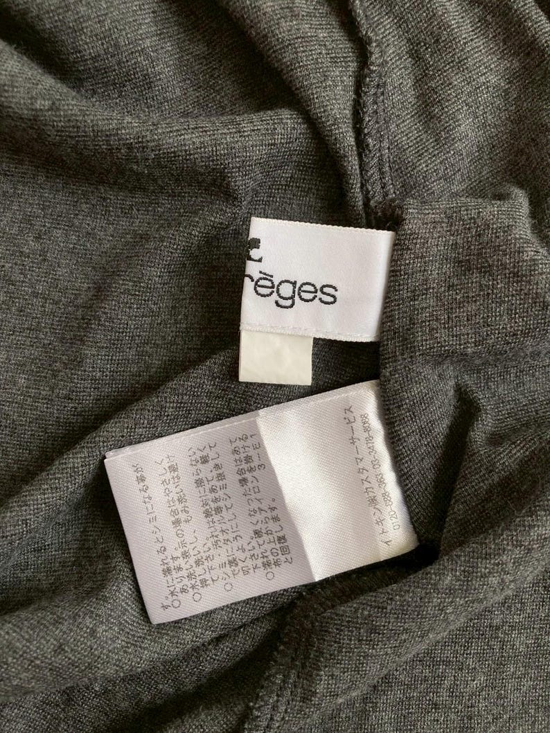 Puede incluir: Primer plano de una tela gris oscuro con dos etiquetas blancas. Una etiqueta tiene el nombre de la marca "r&egrave;ges" impreso. La otra etiqueta contiene texto japon&eacute;s con instrucciones de cuidado. La tela parece ser un material de punto.