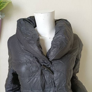 May include: A dark grey quilted puffer coat with a large, draped collar and toggle fastenings. The coat has a slightly shiny finish. The mannequin's head is white.