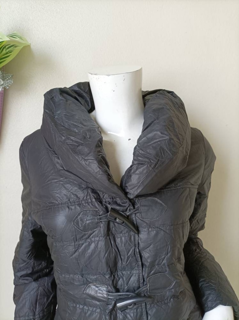 May include: A dark grey quilted puffer coat with a large, draped collar and toggle fastenings. The coat has a slightly shiny finish. The mannequin's head is white.