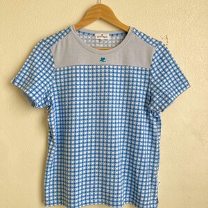 Może przedstawiać: T-shirt z kr&oacute;tkim rękawem w niebiesko-białą kratę. G&oacute;ra koszulki jest szara, z nazwą marki i logo. Koszulka wisi na drewnianym wieszaku.