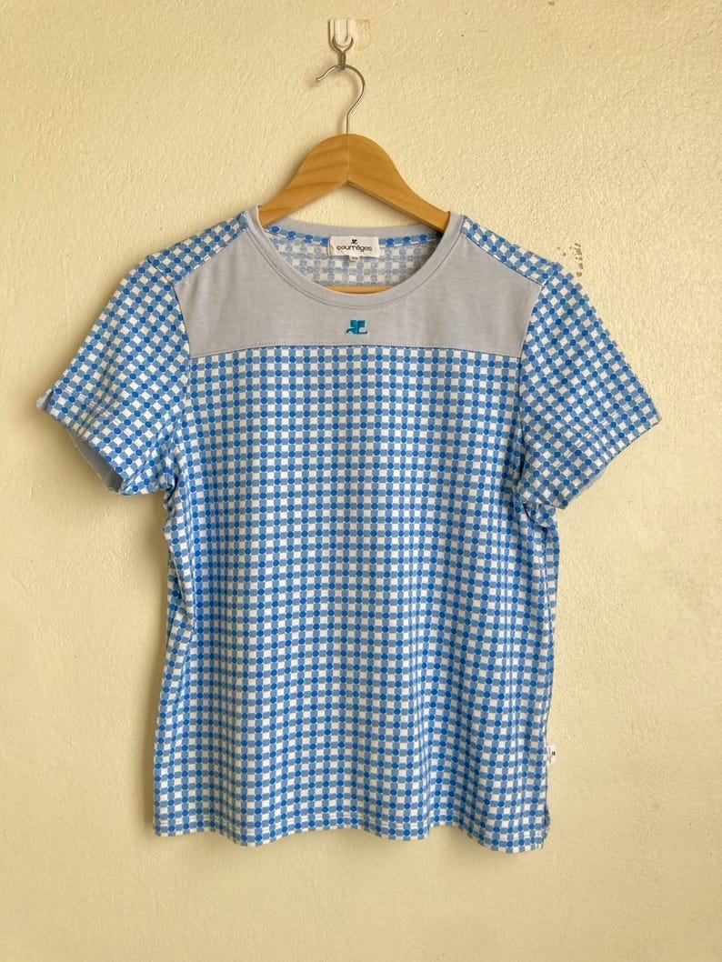 Może przedstawiać: T-shirt z kr&oacute;tkim rękawem w niebiesko-białą kratę. G&oacute;ra koszulki jest szara, z nazwą marki i logo. Koszulka wisi na drewnianym wieszaku.