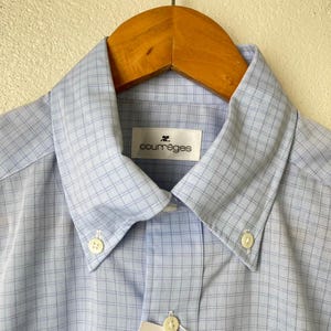May include: A light blue button-down shirt with a white and light blue checkered pattern. The shirt has a collar and a button closure. The brand name "Courr&egrave;ges" is printed on a white tag attached to the collar.