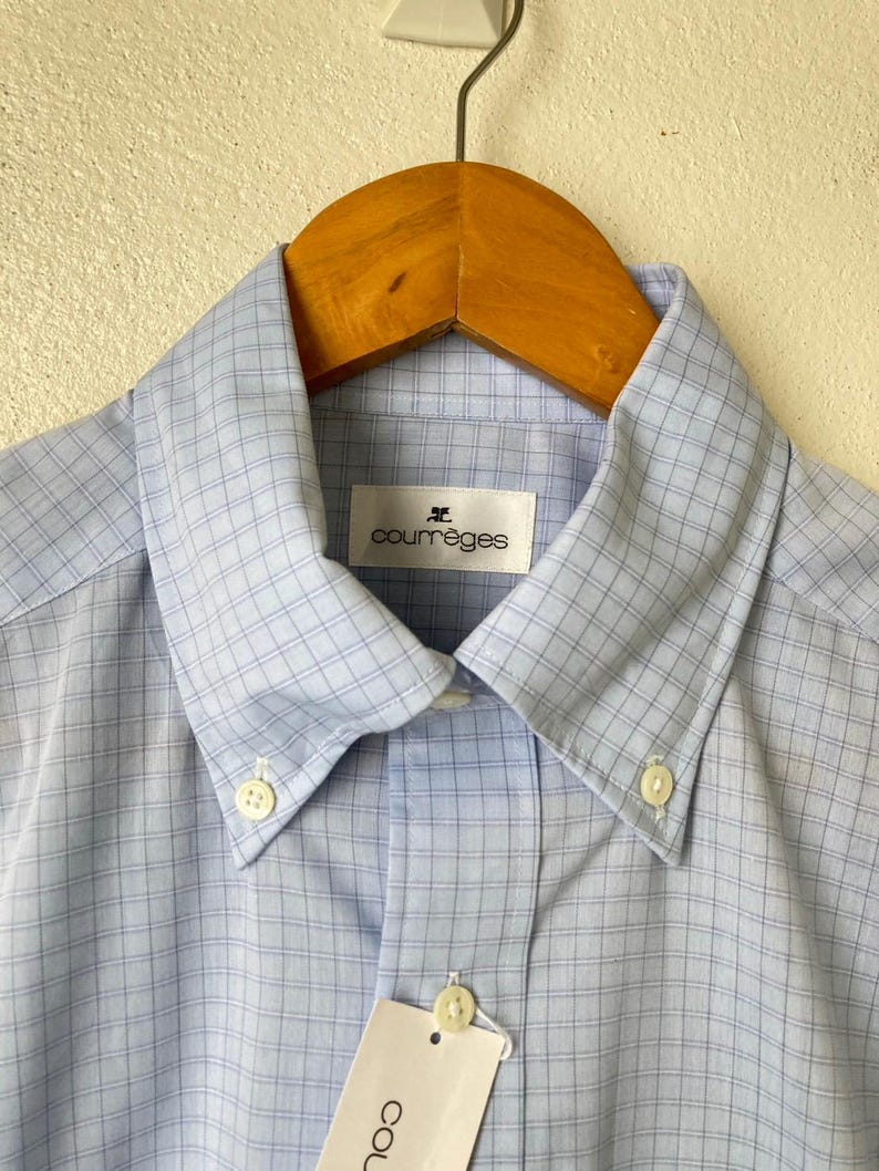 May include: A light blue button-down shirt with a white and light blue checkered pattern. The shirt has a collar and a button closure. The brand name "Courr&egrave;ges" is printed on a white tag attached to the collar.