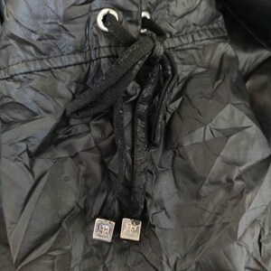 May include: Close-up of a black drawstring closure on a garment. The drawstring is black and knotted, with silver-coloured metal eyelets and square end pieces. The fabric is a dark, textured material.