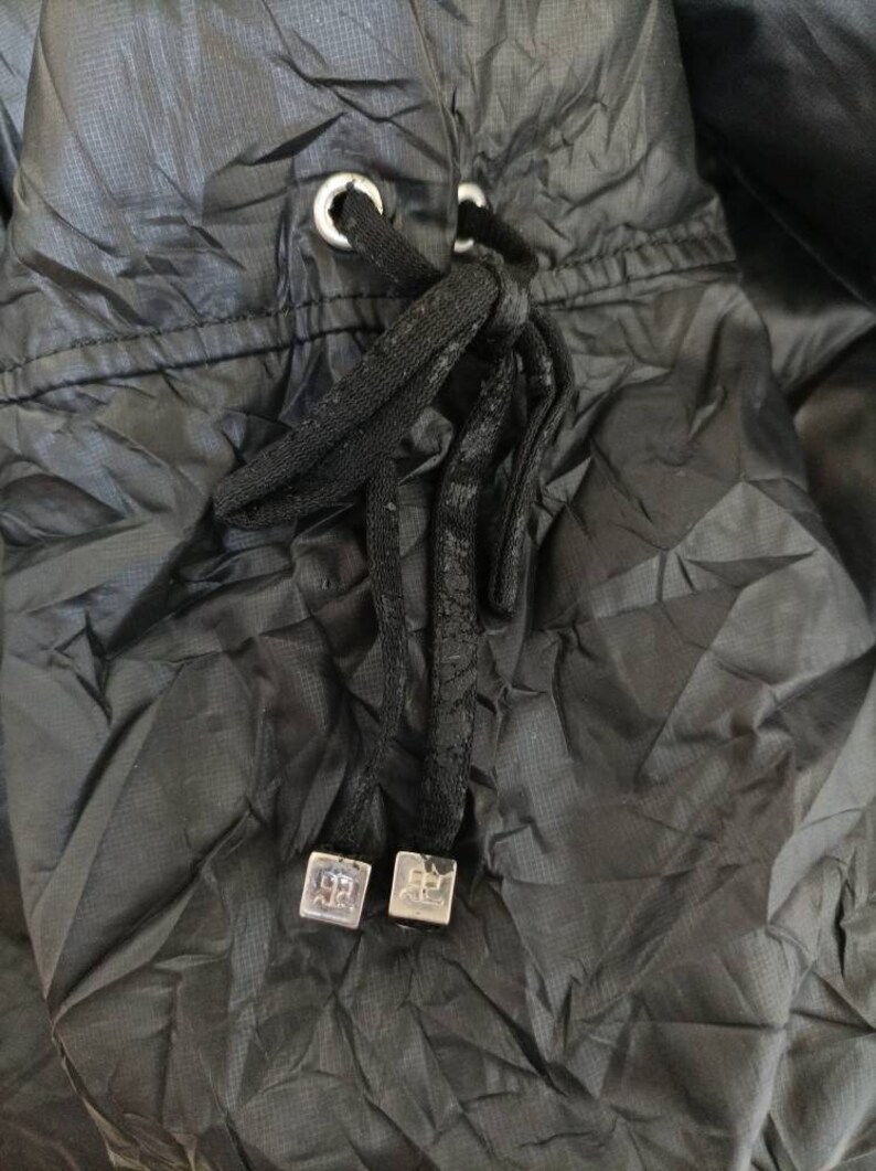 May include: Close-up of a black drawstring closure on a garment. The drawstring is black and knotted, with silver-coloured metal eyelets and square end pieces. The fabric is a dark, textured material.