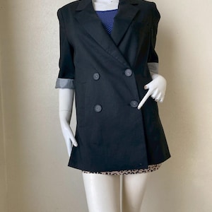 Pode incluir: Um blazer preto de abotoamento duplo com mangas três quartos. As mangas são dobradas, revelando um padrão xadrez preto e branco. O blazer tem quatro botões pretos e é exibido em um manequim.