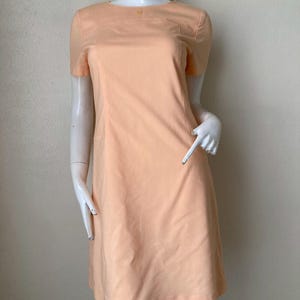 K&ouml;nnte beinhalten: Pfirsichfarbenes Kleid mit kurzen &Auml;rmeln und rundem Ausschnitt. Das Kleid hat ein Knopfdetail am Ausschnitt und eine taillierte Passform. Das Kleid wird auf einer Schaufensterpuppe pr&auml;sentiert.